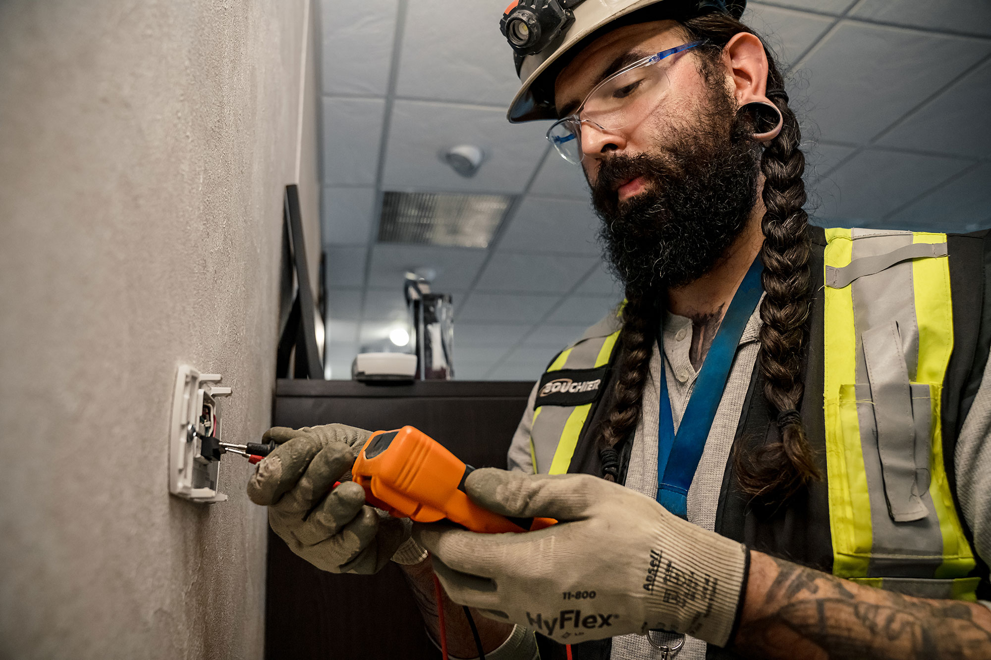 An electrician wearing safety gear, including a hard hat, gloves, and reflective vest, uses a voltage tester on a wall outlet inside a commercial building.