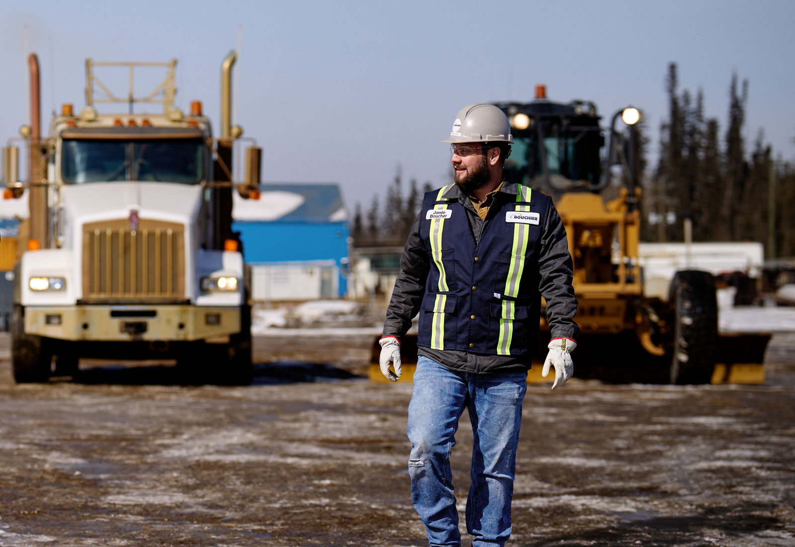 Bouchier worker in a hard hat and safety vest walking confidently on a muddy industrial site, with a transport truck and grader in the background.