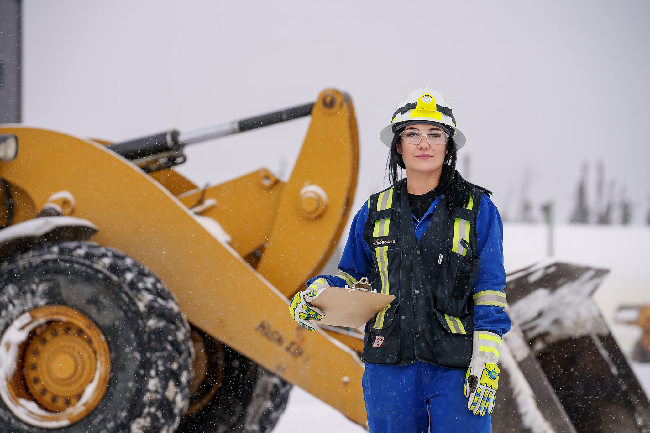 Bouchier team member in full safety gear holding a clipboard, standing in front of heavy equipment during snowy weather on an industrial job site.