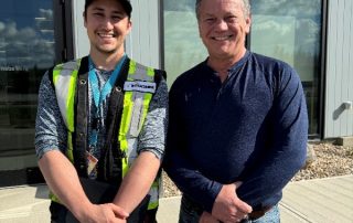 Evan standing outside a Bouchier facility with a colleague, wearing a high-visibility vest and holding a tablet, both smiling on a sunny day.