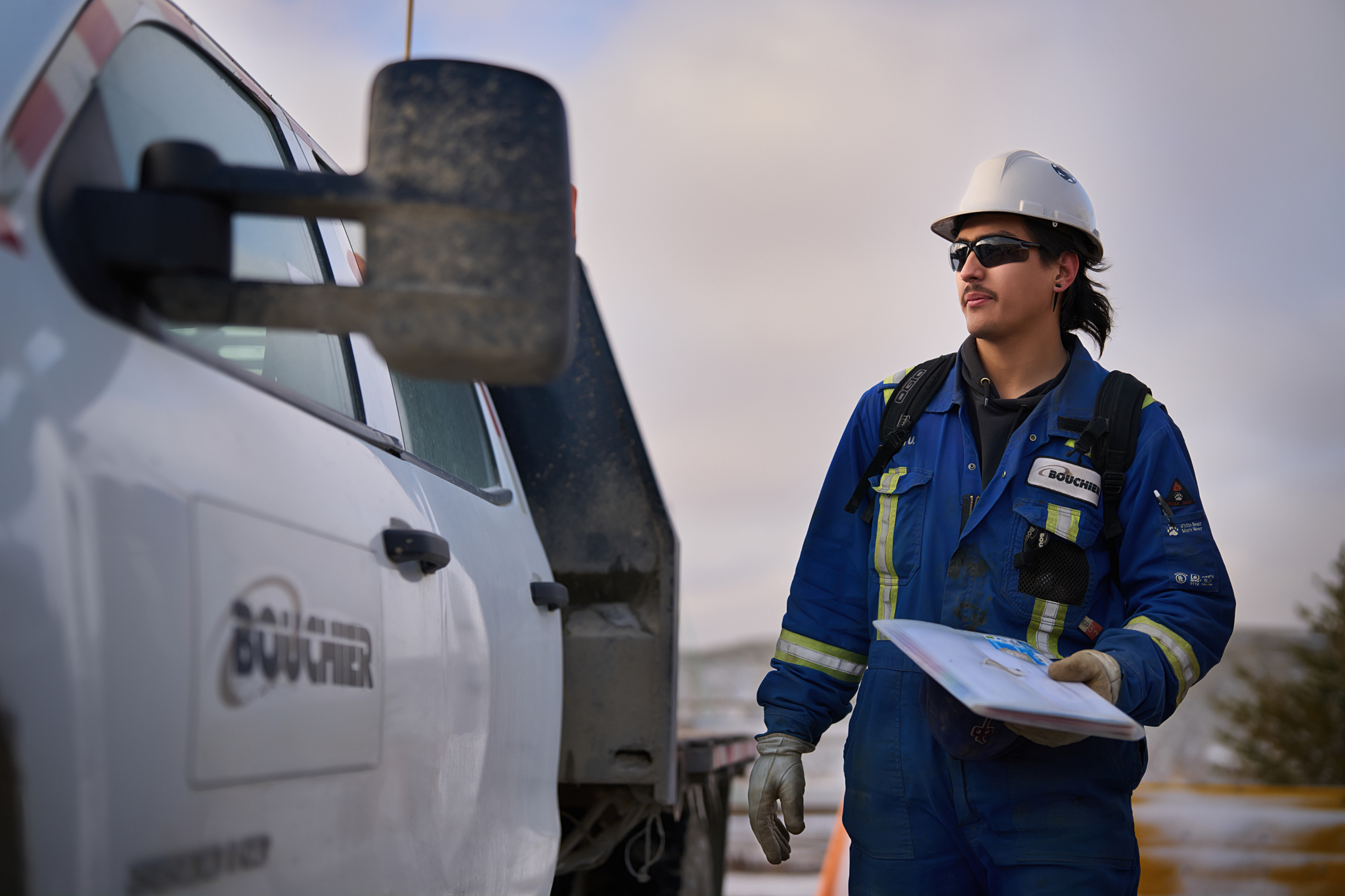 Bouchier team member in blue coveralls and a hard hat holding site documents, standing beside a work truck on an industrial job site.