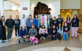Group photo of students and staff posing indoors with a framed pink sports jersey and a dog wearing a matching pink outfit. The group stands in front of a wall-mounted buffalo hide in a bright, modern building.