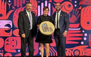 Three people standing on stage in front of a vibrant red and purple Indigenous art backdrop. The woman in the center holds a hand-painted drum featuring traditional artwork.