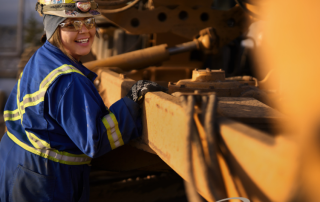 Smiling construction worker wearing safety gear and a sticker-covered hard hat, standing beside heavy machinery with the Bouchier logo visible in the corner.